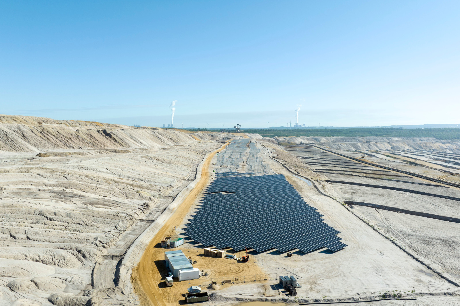 Ein Solarpark in einer kahlen Landschaft, mit blauem Himmel und in der Ferne sichtbaren Kraftwerken.