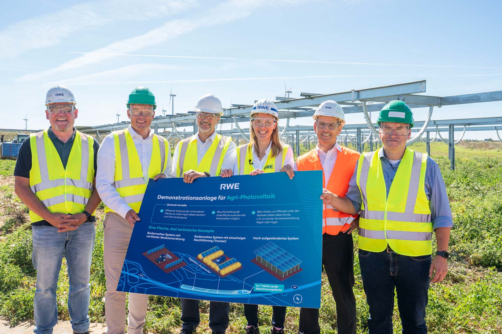 Fünf Personen in Sicherheitswesten und Helmen präsentieren ein Info-Plakat über Agri-Photovoltaik in einem Sonnenfeld.