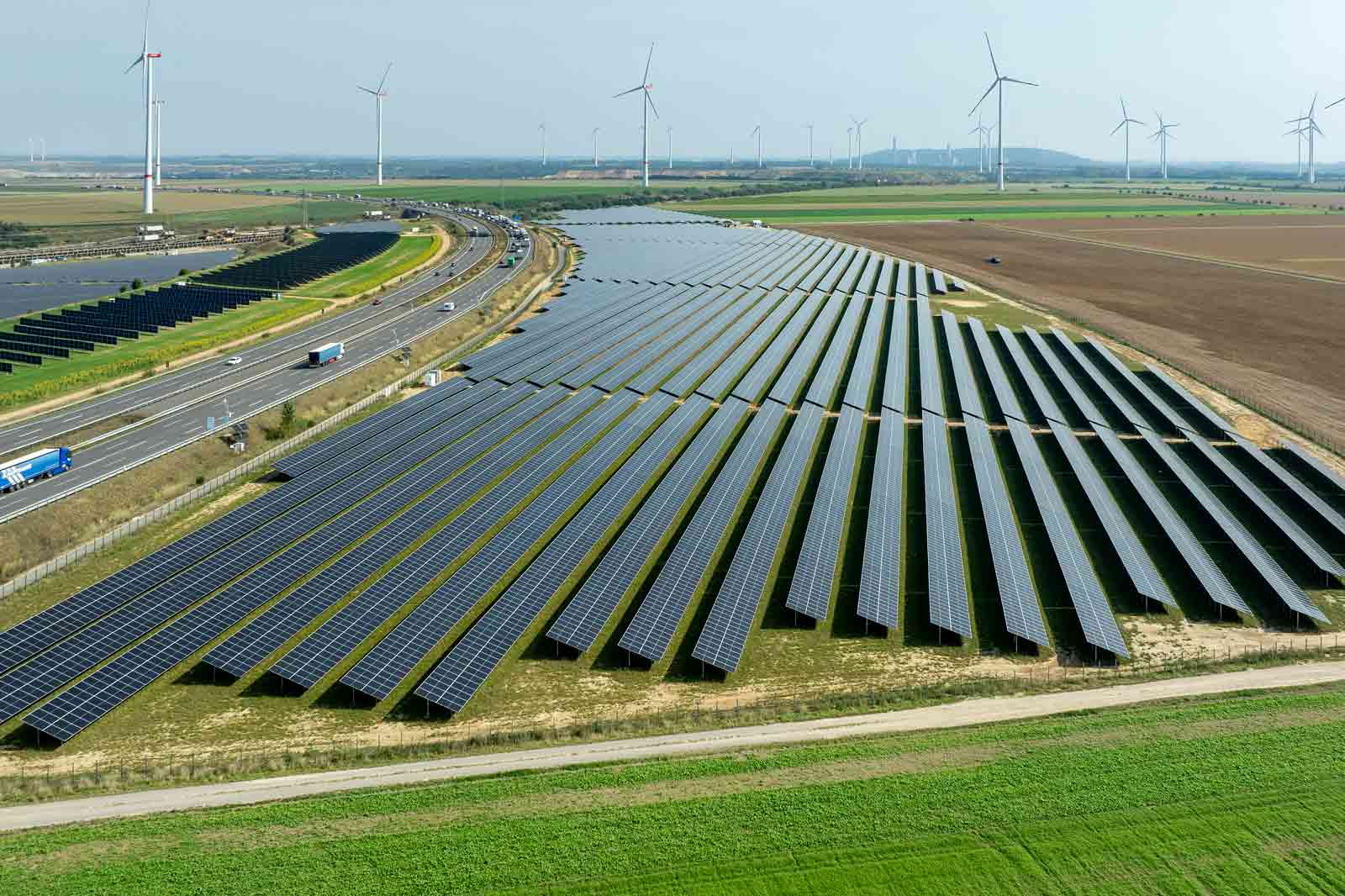 Vogelperspektive auf Solarpanele, die in Reihen neben einer Autobahn angeordnet sind, mit Windkraftanlagen im Hintergrund.