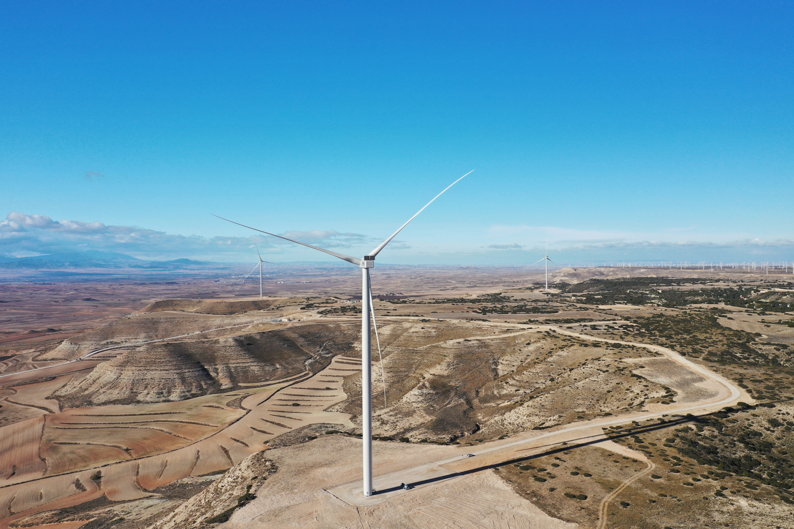 Eine Windkraftanlage steht auf hügeligem Gelände mit sanften Erdwellen und einem klaren, blauen Himmel im Hintergrund.