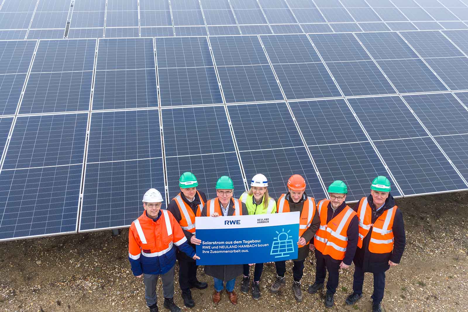 Gruppenfoto von Arbeitern mit Sicherheitswesten und Helmen vor Solarpanels, halten ein Schild mit der Aufschrift 'Solarstrom aus dem Tagebau'.