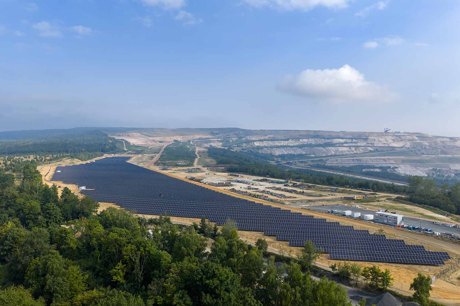 Eine weite Ansicht eines Solarparks mit vielen Solarpanelen, umgeben von Bäumen und sanften Hügeln im Hintergrund.