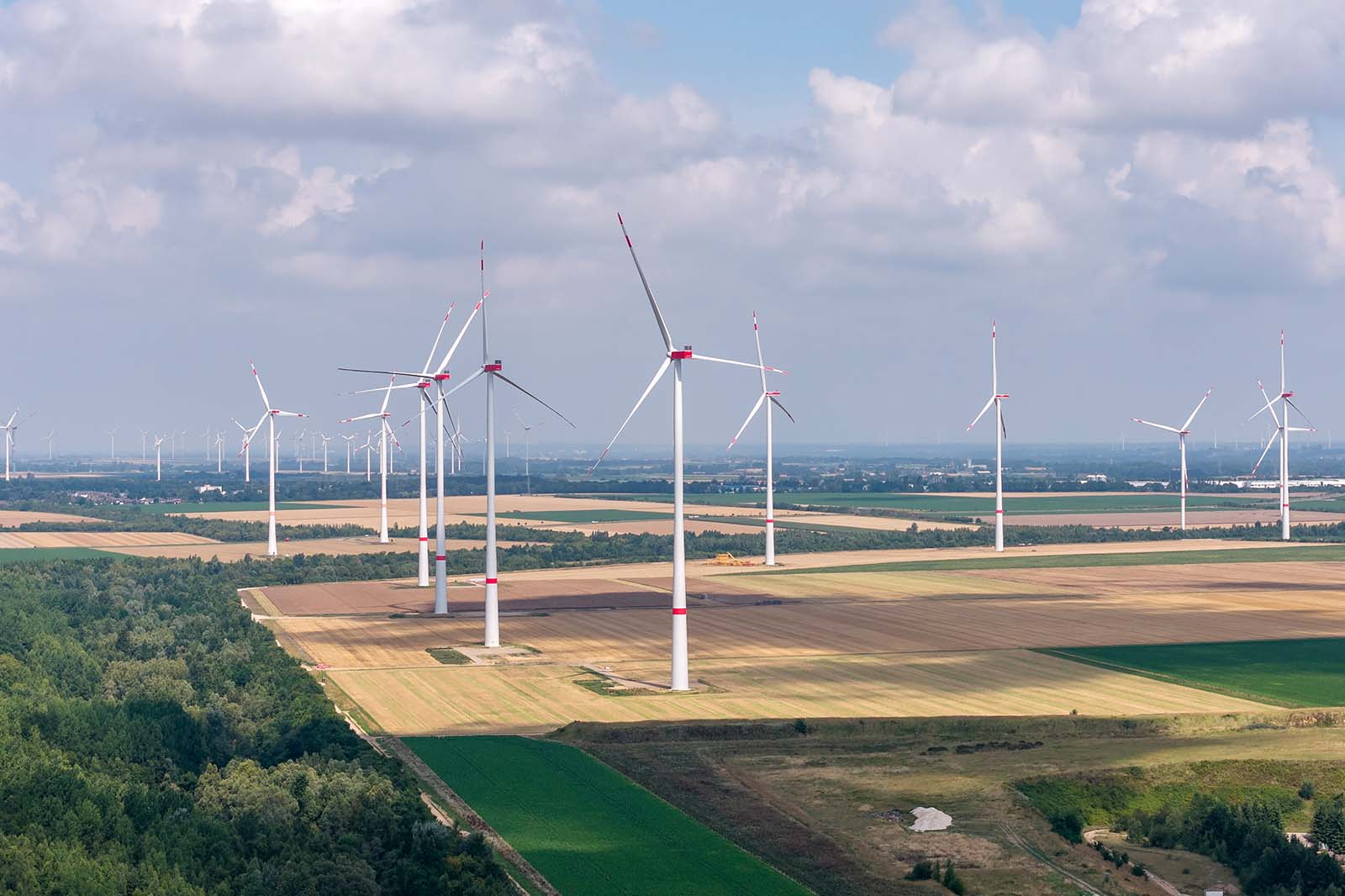 Luftaufnahme von Windrädern über Feldern unter wolkigem Himmel, mit Baumstreifen, Schatten und roten Markierungen an Rotorblättern.