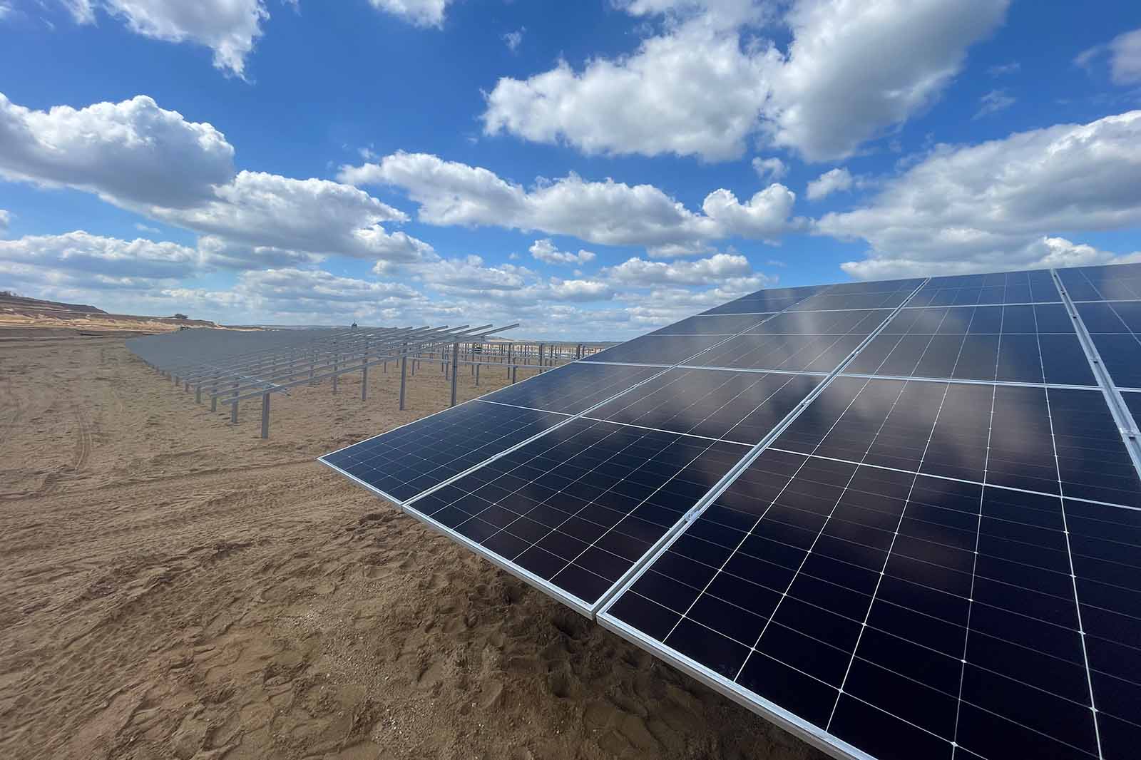 Glänzende dunkelblaue Solarmodule mit silbernen Rahmen dominieren den Vordergrund auf sandigem Boden, Reihen ziehen sich unter einem strahlend blauen Himmel mit weißen Wolken.