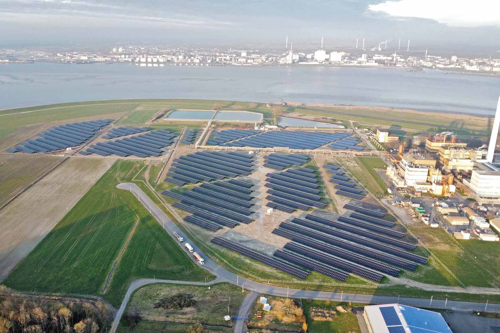 Luftaufnahme weitläufiger blauer Solarmodulfelder auf flachem, grünen Küstenland neben beigen Industriegebäuden und ruhigem Hafenwasser im Hintergrund.