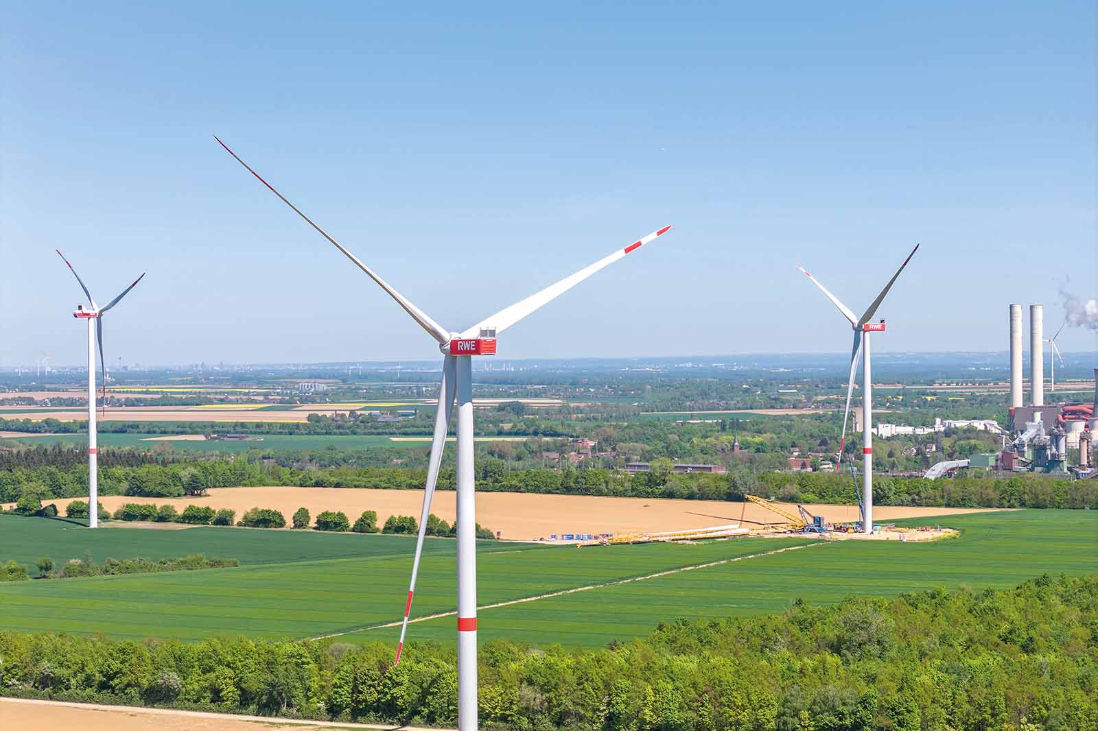 Drei weiße Windräder aus Metall mit roten Markierungen stehen über grünen Feldern vor klarem blauem Himmel. Die Szene wirkt ruhig und weit.