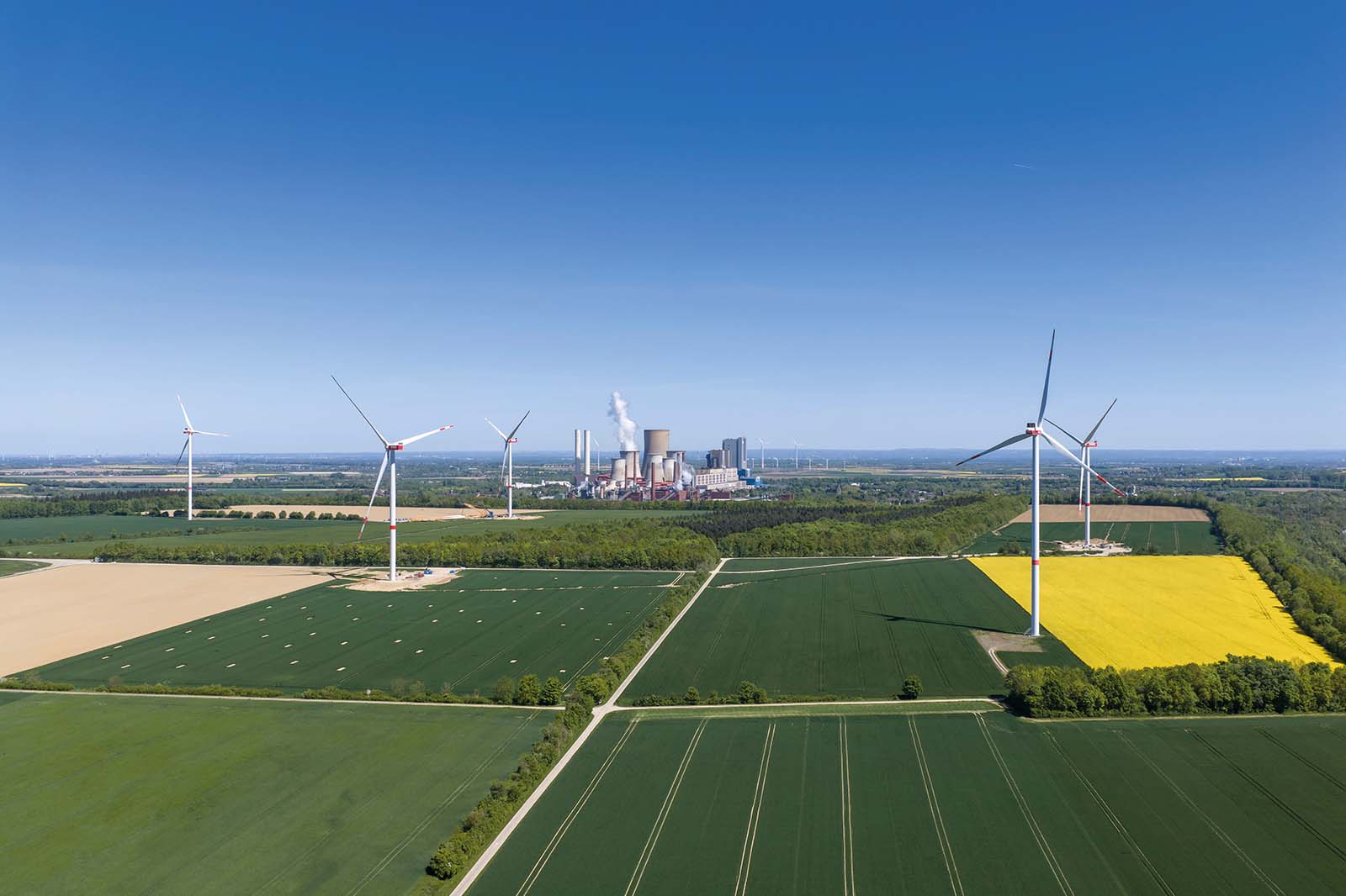 Weiße Windräder stehen in der Mitte grüner und gelber Felder, im Hintergrund ein Kraftwerk mit rauchenden Schloten unter klarem blauem Himmel.