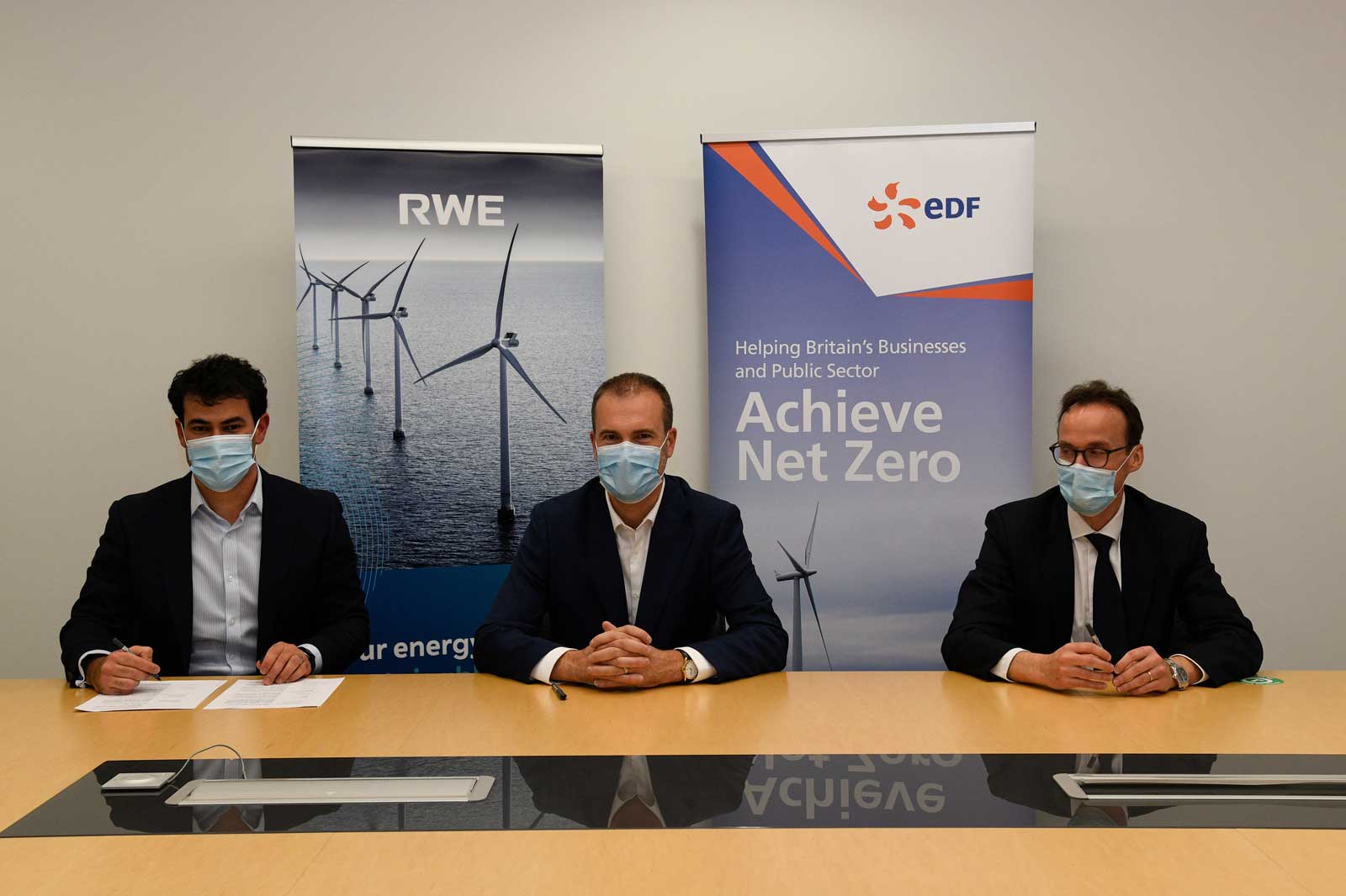 Drei Personen sitzen an einem Tisch. Im Hintergrund sind Banner von RWE und EDF zu sehen, die Windkraft symbolisieren.