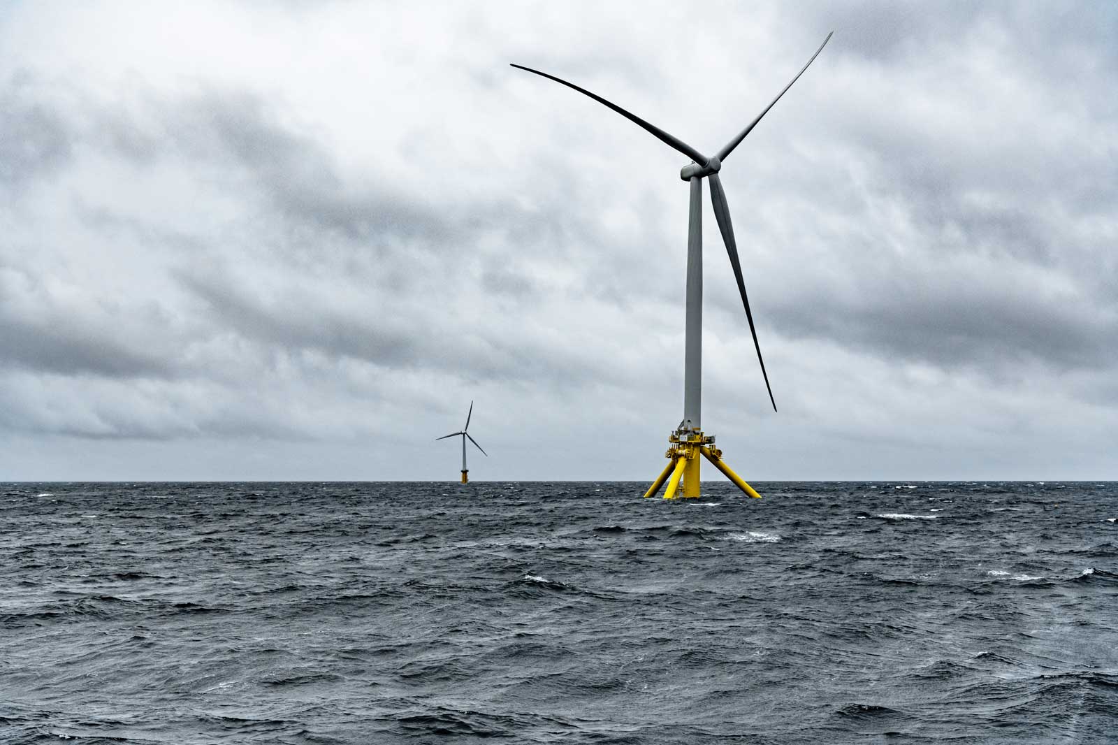 Zwei Offshore-Windkraftanlagen stehen in stürmischer See unter einem bewölkten Himmel.