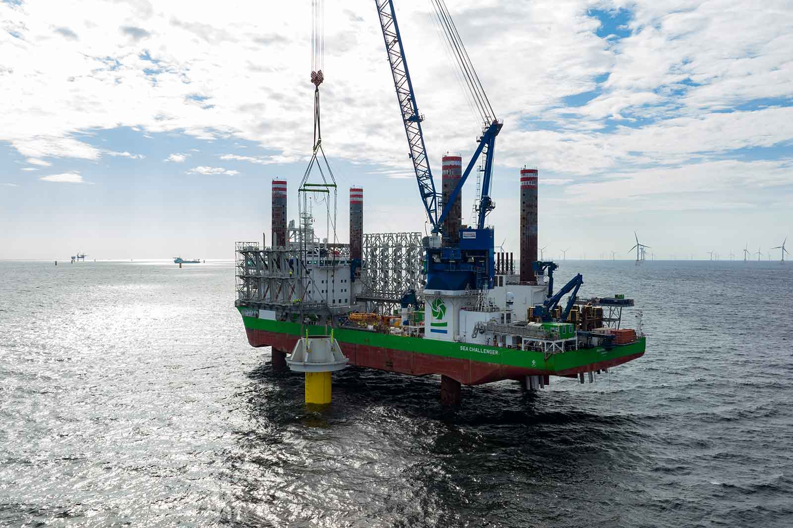 Ein Arbeitsboot mit Kränen installiert eine Struktur im Meer. Windkraftanlagen sind im Hintergrund sichtbar.
