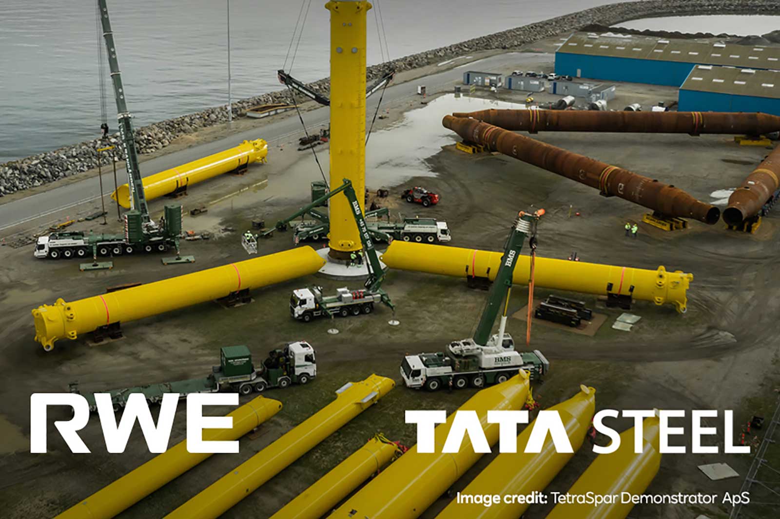 An aerial view of a construction site with yellow cylindrical structures, cranes, and trucks, indicating offshore wind turbine preparation.