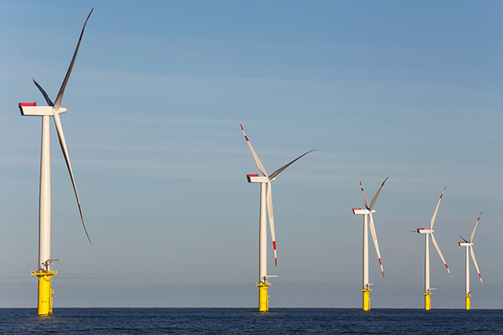 Eine Reihe von Windkraftanlagen mit gelben Fundamenten steht im Wasser unter einem klaren Himmel.