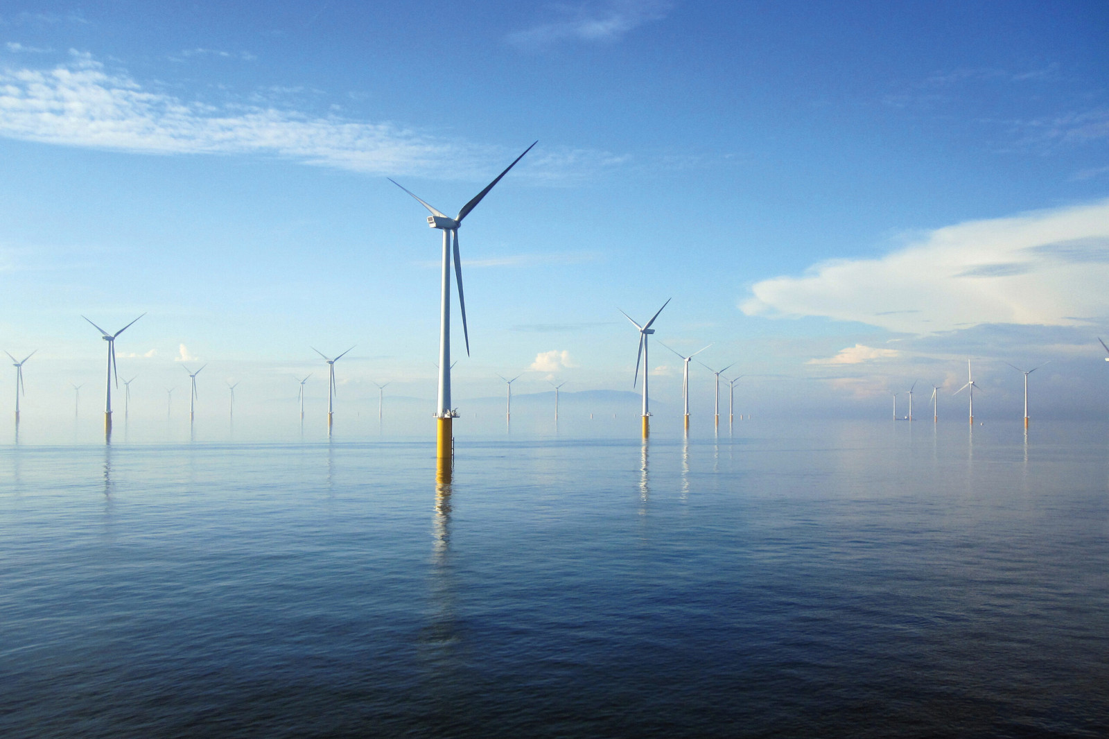 Eine Reihe von Offshore-Windturbinen steht in ruhigen Gewässern unter einem klaren blauen Himmel mit einigen Wolken.