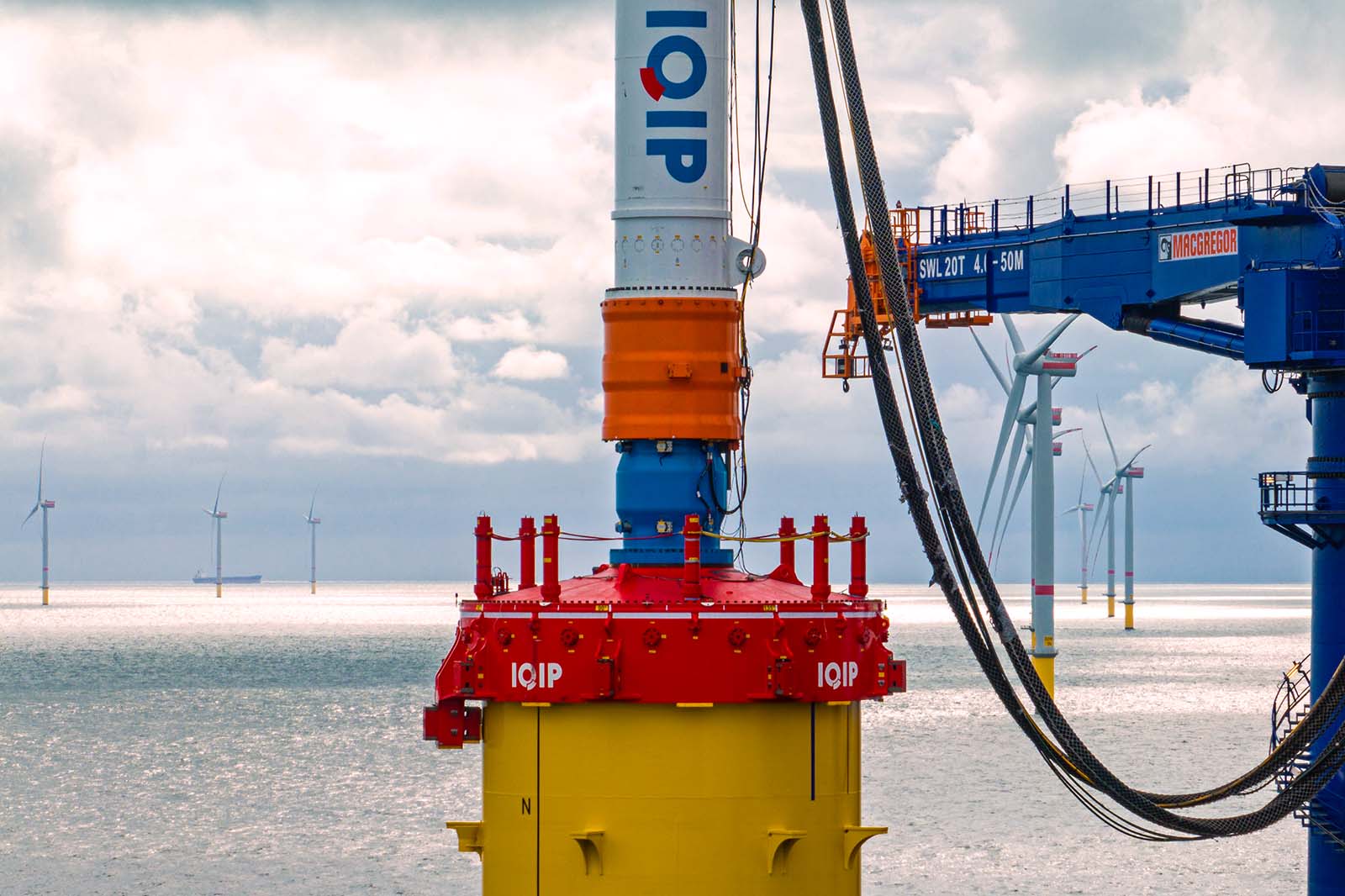 Ein Kran installiert eine rot-gelbe Offshore-Windturbinen-Basis mit dicken Kabeln, und mehrere Windturbinen stehen am Horizont unter wolkigem Himmel.
