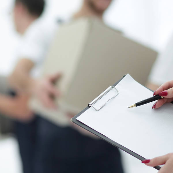 Eine Person hält ein Notizblatt mit einem Stift, während im Hintergrund ein anderer Mensch eine Kiste trägt.