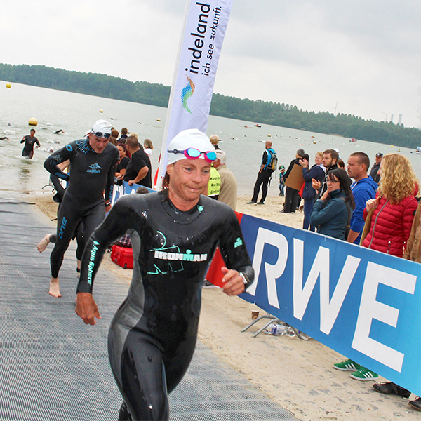 Swimmers in wetsuits exit the water at a competition, surrounded by spectators and a promotional banner.