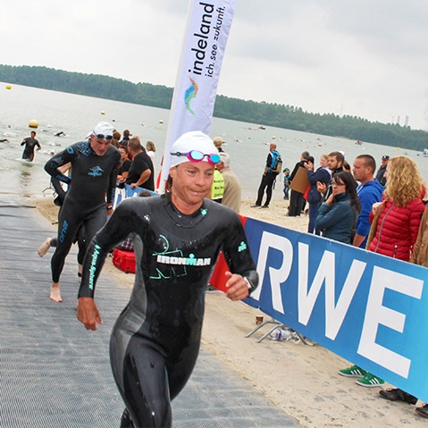 Swimmers in wetsuits exit the water at a competition, surrounded by spectators and a promotional banner.