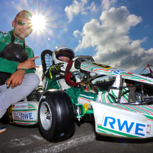 A kart driver poses with their kart at the racetrack, wearing a helmet while the driver points with their finger.