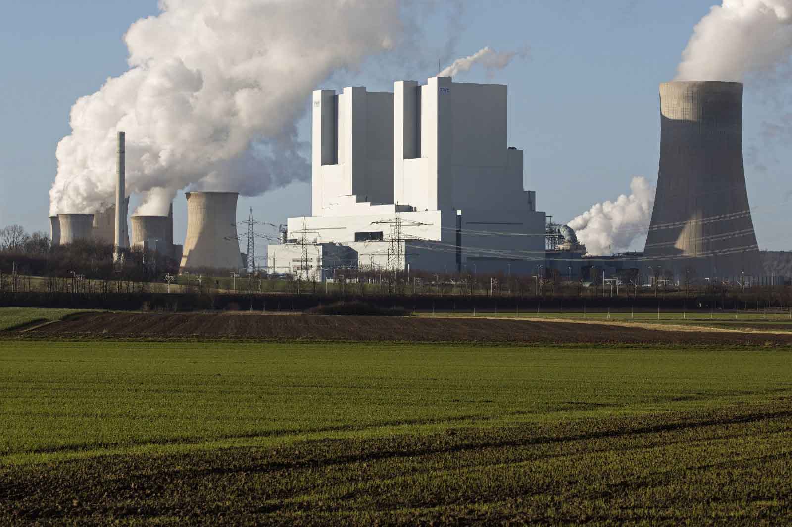 Ein Kraftwerk mit Kühltürmen stößt Dampf in den klaren Himmel aus und ist von Feldern umgeben.