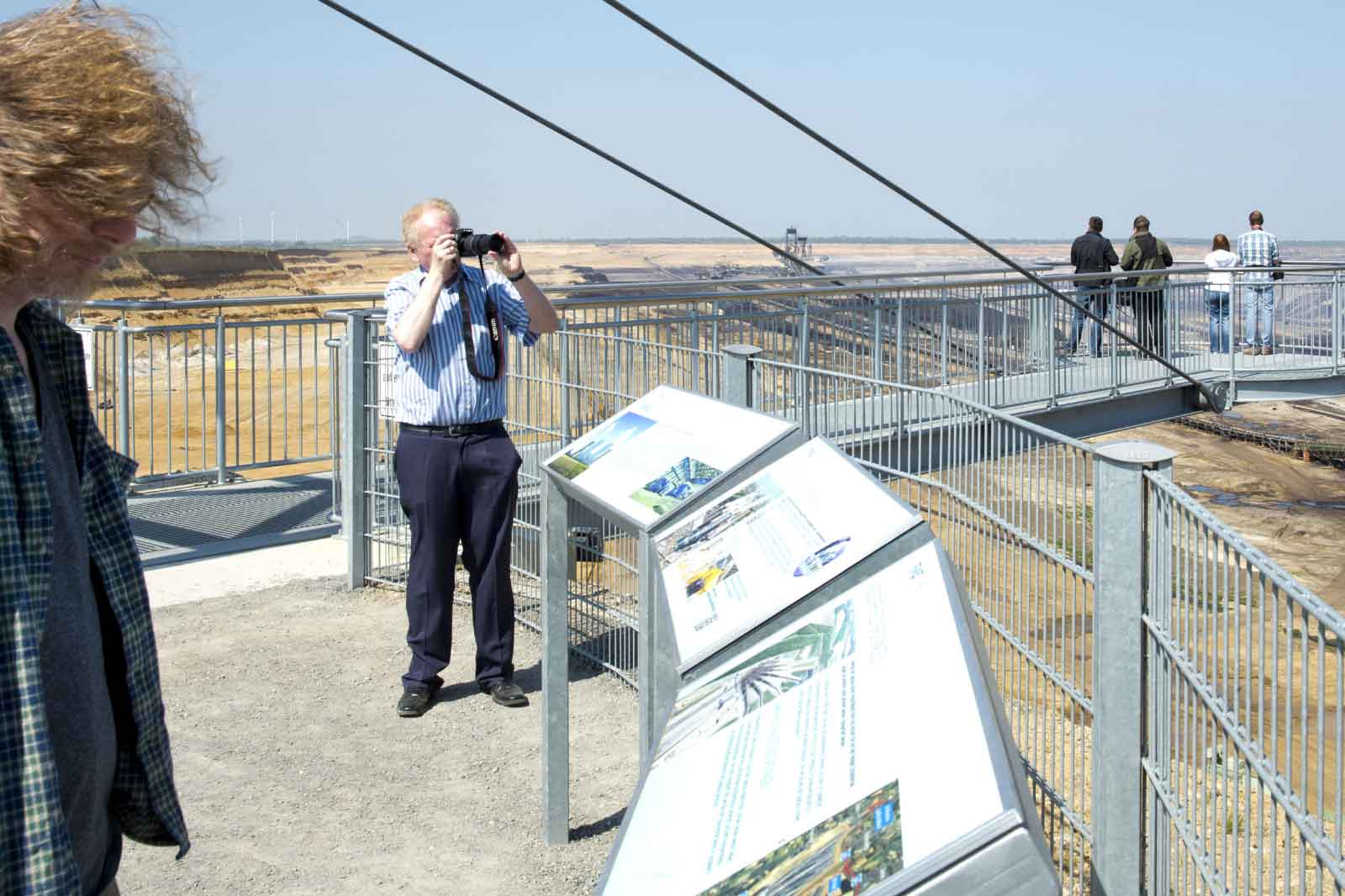 Besucher betrachten eine große Tagebau-Landschaft von einer Aussichtsplattform mit informativen Schildern.