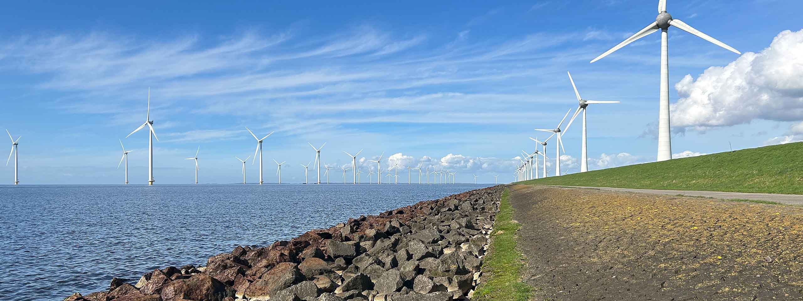 Panoramic view of an offshore wind farm with turbines in the water and on the grassy dyke, a rocky shoreline and a blue sky.