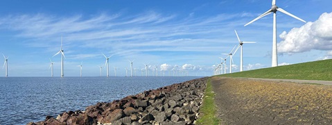 Panoramic view of an offshore wind farm with turbines in the water and on the grassy dyke, a rocky shoreline and a blue sky.