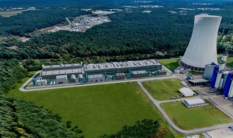 Aerial view of a large industrial facility surrounded by a forested area, featuring cooling towers and grey buildings.