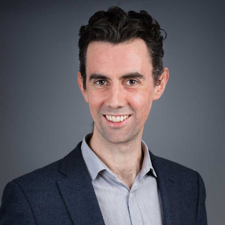 Ein Mann in einem dunkelblauen Blazer mit einem grauen Hemd vor einem neutralen Hintergrund.