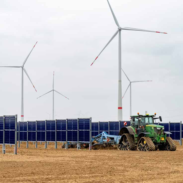 Ein Traktor bearbeitet den Boden zwischen Solarpanelen, während Windkraftanlagen im Hintergrund sichtbar sind.