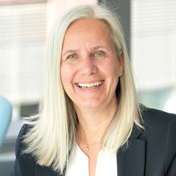 Eine Person mit langen, blonden Haaren trägt einen Blazer. Der Hintergrund zeigt verschwommene Fenster mit natürlichem Licht. Portrait von Nicola Moos.