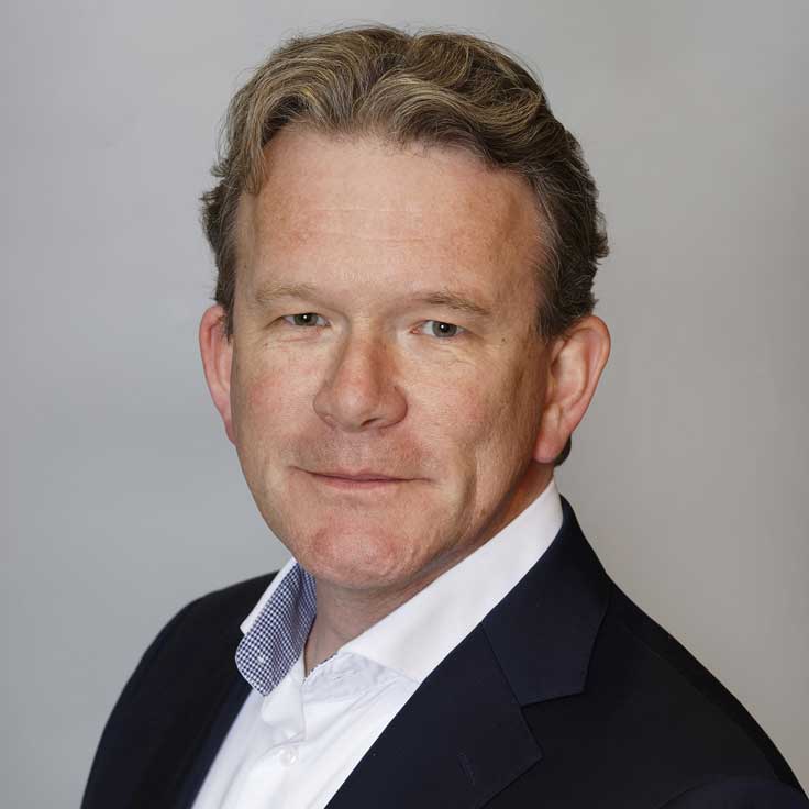 Ein Mann in einem Anzug mit blonden Haaren, trägt ein weißes Hemd und einen blauen Blazer, vor einem grauen Hintergrund.