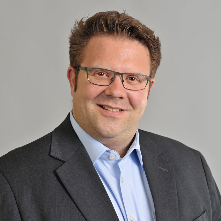 Eine Person in einem dunklen Anzug mit hellblauer Hemd auf einem grauen Hintergrund. Portrait von Jan Peter Cirkel.
