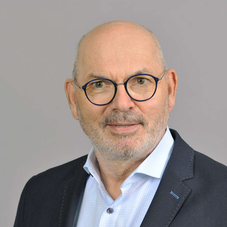 Ein Mann mit einem dunklen Jacket und einem hellblauen Hemd steht vor einem grauen Hintergrund. Portrait von Lothar Lambertz.