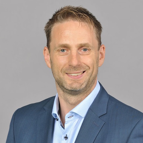 Ein Mann trägt einen blauen Anzug und ein hellblaues Hemd vor einem grauen Hintergrund. Portrait von Matthias Beigel.
