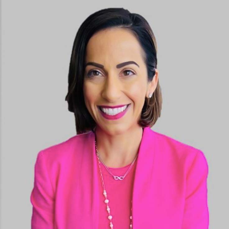 Eine Person trägt einen pinken Blazer und eine Kette. Der Hintergrund ist grau. Portrait von Patricia Kakridas.