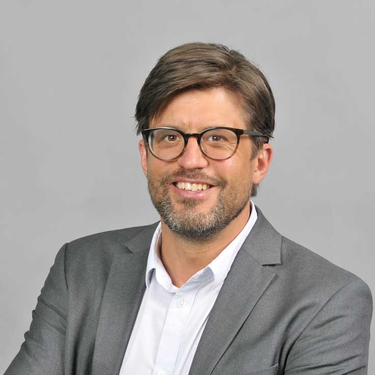 Ein Mann in einem grauen Anzug mit einem weißen Hemd, der in Richtung Kamera schaut und seine Arme verschränkt. Portrait von Simon Lorenz.