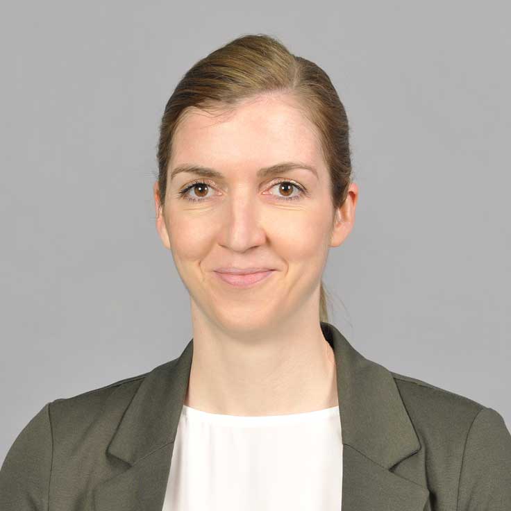 Eine professionelle Frau in einem dunklen Blazer und einer weißen Bluse vor einem neutralen grauen Hintergrund. Portrait von Sinje Vogelsang.