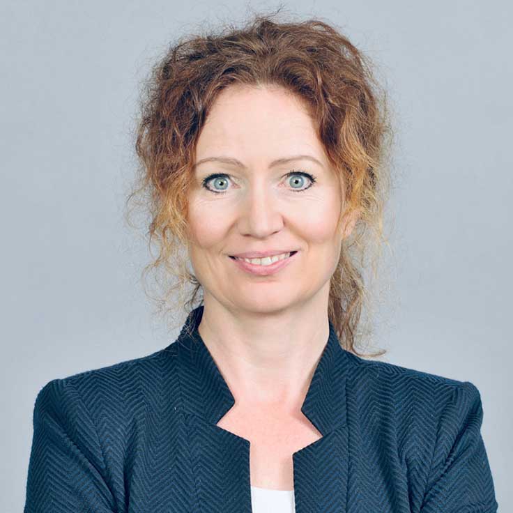 Eine Person mit lockigem, rotem Haar trägt ein schwarzes, strukturiertes Jackett und hat die Arme gekreuzt. Portrait von Vera Bücker.