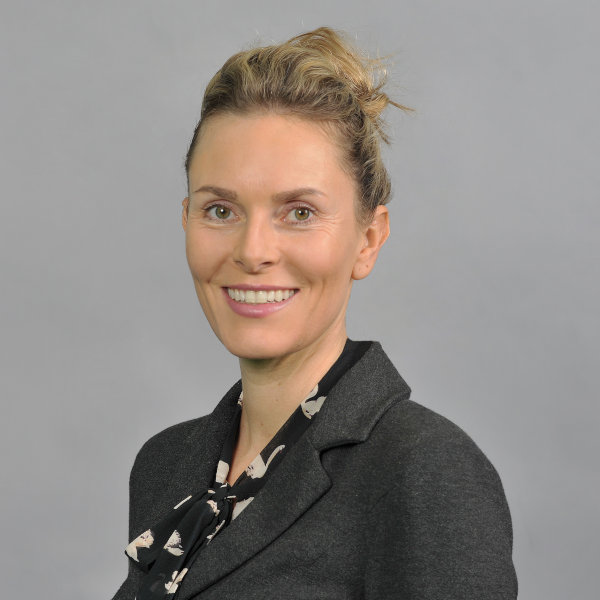Eine Person in einem schwarzen Blazer mit einem gemusterten Schal, auf einem grauen Hintergrund stehend. Portrait von Viola Baumann.