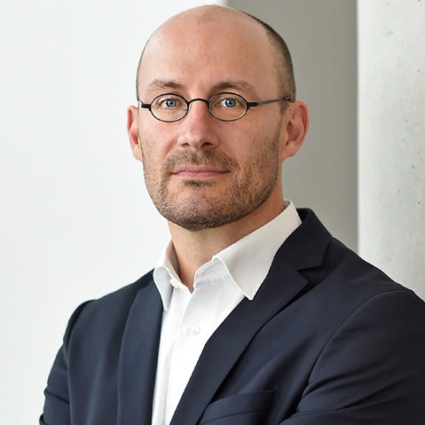 Ein Mann in einem dunkelblauen Anzug mit einem weißen Hemd steht vor einer hellen Wand und sieht professionell aus. Portrait von Olaf Winter.