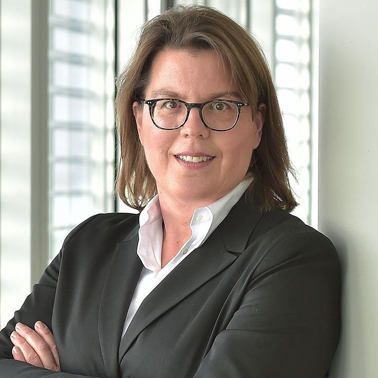 Eine Person in einem schwarzen Anzug steht mit verschränkten Armen vor einem modernen Fenster mit vielen Lichtern. Portrait von Regina Wolter.