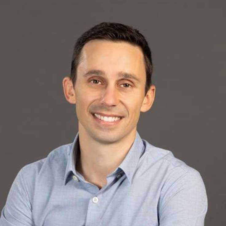 A portrait of a man wearing a light blue shirt. The background is grey, and he is posing in a relaxed manner. Portrait of Andrea Baldi.