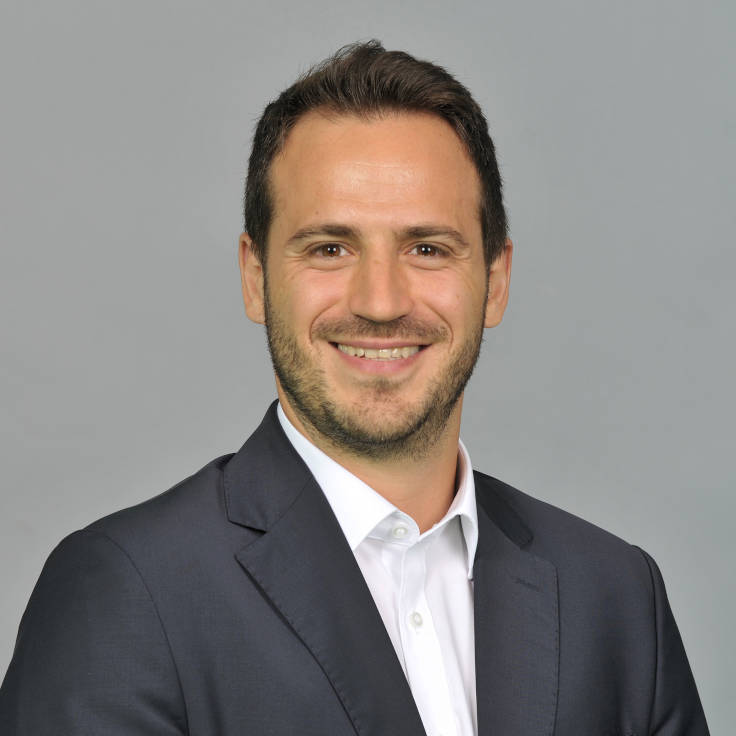 A man wearing a dark suit and white shirt poses against a grey background. Portrait of Cihan Sönmez.