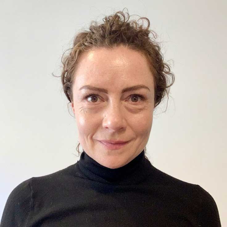 A person with curly hair is wearing a black jumper. The background is bright and neutral. Portrait of Erika Vasquez.