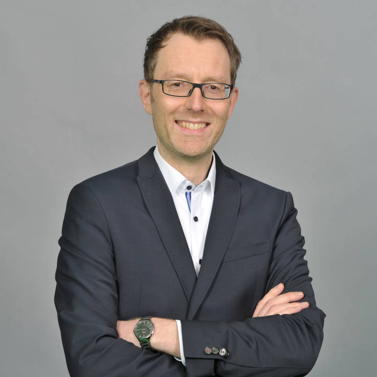 A man in a suit stands with his arms folded in front of a gray background and radiates professionalism. Portrait of Kai Stevens.