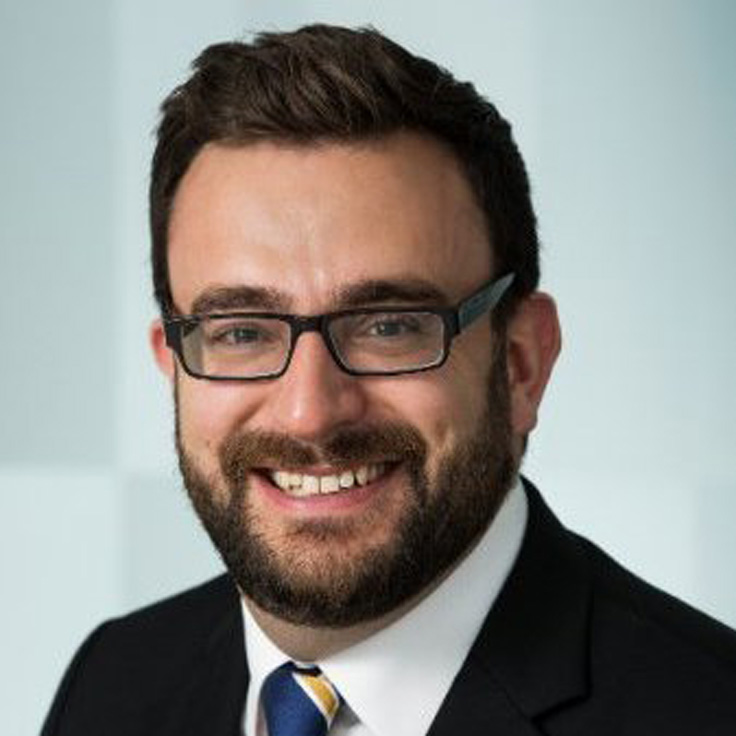 A person with short dark hair in a black suit jacket, white shirt and blue-yellow striped tie against a pale gray background. Portrait of Nick Stapley.