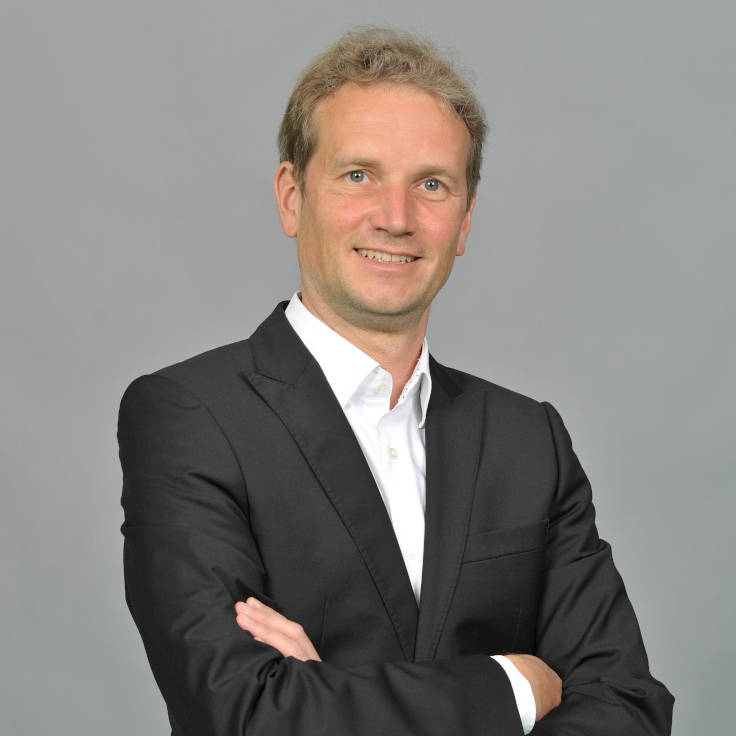 A person in a black suit with folded arms standing in front of a gray background. Portrait of Nils Weise.