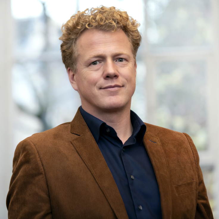 A man with curly blonde hair in a brown cord jacket and a navy shirt stands in front of large windows. Portrait of Peter den Breejen.