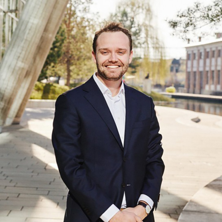 A man in a dark blue suit and white shirt stands on a brightly paved promenade in front of modern columns and a water canal. Portrait of Pim de Jong.