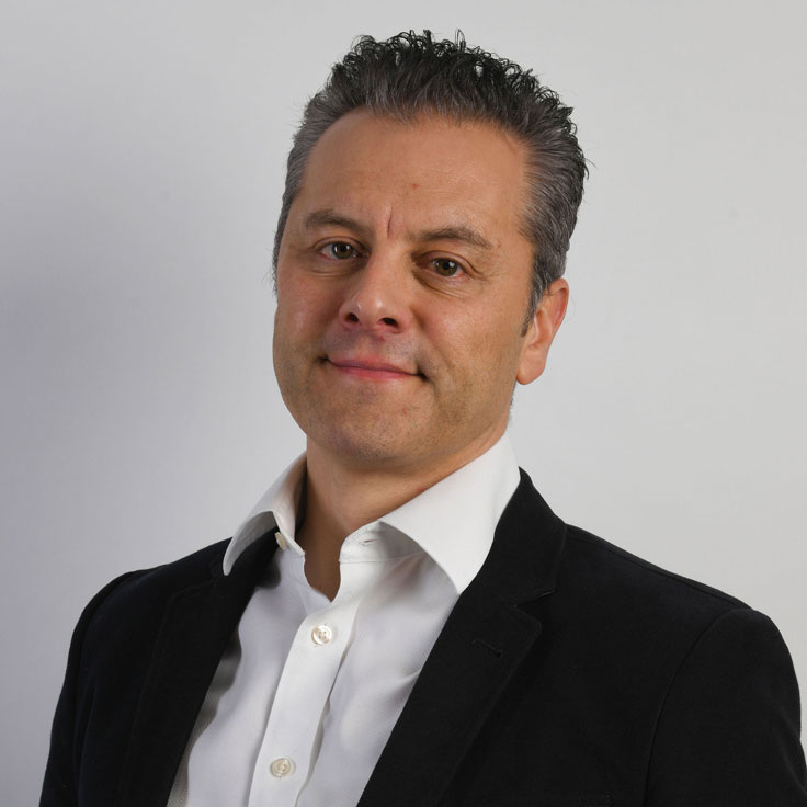 A head-and-shoulders portrait of a person with short dark hair wearing a white button-up shirt and a black blazer against a light gray background. Portrait of Richard Liversidge.